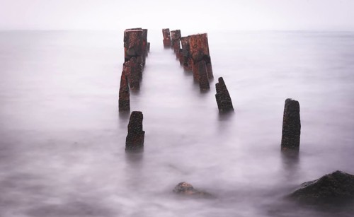 Wooden bridge remains in silky water with sea horizon in the background. Concept of time and
