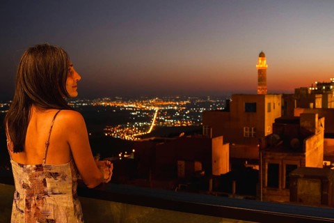 Tourist woman on terrace restaurant balcony watching illuminated skyline of Mardin with glowing