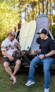 Two male friends talking sitting on a truck with their dog and surfboards