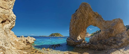 Stone arch on Grande Blue Beach in front of a small island and a rocky coastline under a clear blue