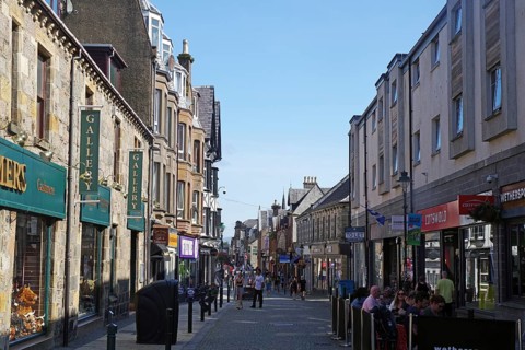Busy shopping street with people and traditional buildings under a blue sky, Fort William,