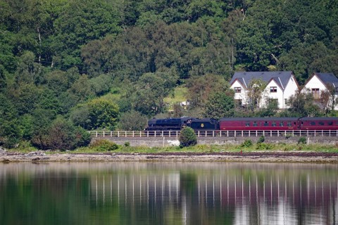 A train travelling along a loch, past houses and a dense green forest, Hogwarts Express, Jacobite