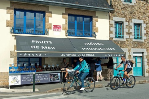 Cyclists on the Quai John Kennedy pass a shop selling seafood such as oysters and mussels, Cancale,