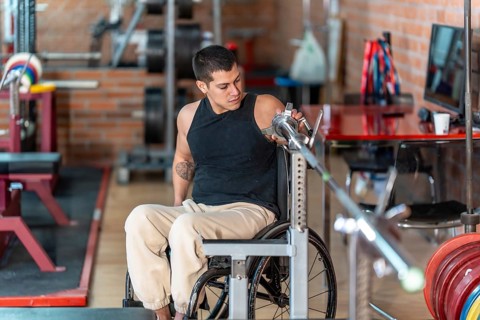 Disabled man in a wheelchair performing weight training with a barbell at a gym, focusing on