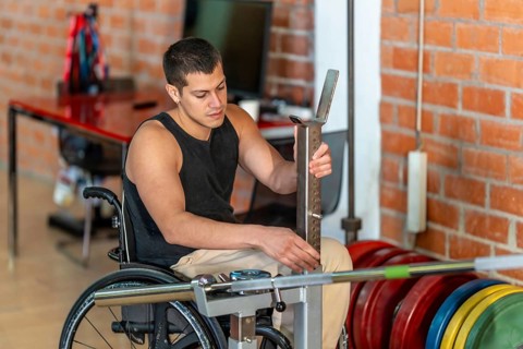 Young man living with a disability sitting in a wheelchair while adjusting a barbell rack during a