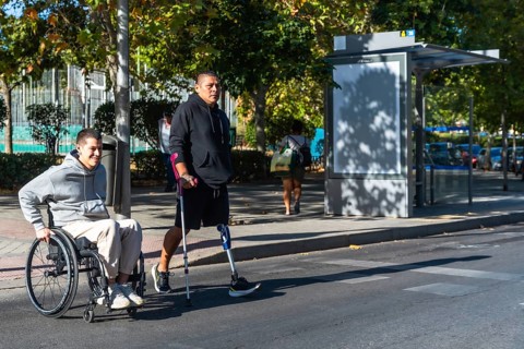 Two amputee men, one using a wheelchair and the other a prosthetic leg with crutch, navigating an