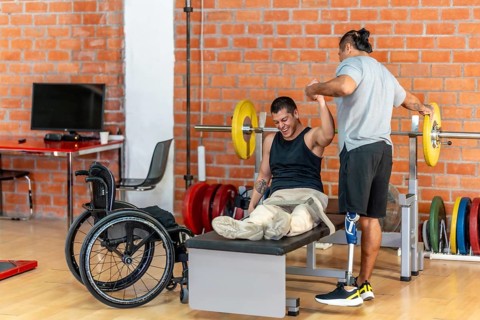Two physically challenged men exercising together, one using a wheelchair for support on a bench