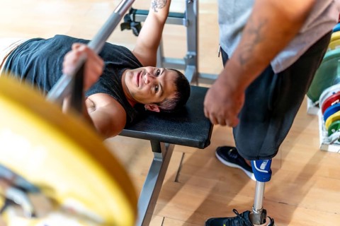 Disabled man performing a bench press with a barbell while a friend with a prosthetic leg stands