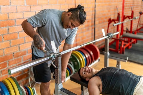 Two diverse men, one with a prosthetic limb assisting the other who is laying on a weight bench,