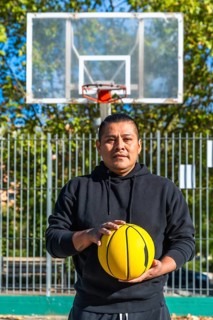 Indigenous man standing on an outdoor basketball court holding a bright yellow basketball, looking