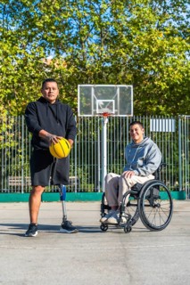 Two men, one with a prosthetic leg holding a basketball and another sitting in a wheelchair,
