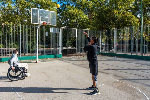 Two athletes playing basketball on an outdoor court. One sitting in a wheelchair and the other
