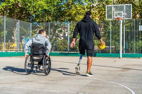 Man with prosthetic leg dribbling basketball on outdoor court while friend in a wheelchair watches,