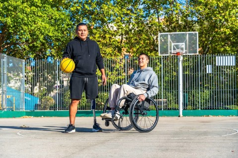 Two young men, one with a prosthetic leg holding a basketball and another sitting in a wheelchair,