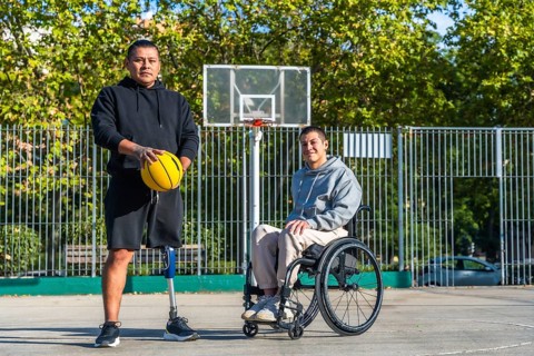 Two young men, one with a prosthetic leg holding a basketball and another sitting in a wheelchair,