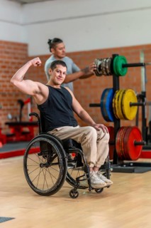 Adaptive athlete in a wheelchair flexing his bicep and smiling while a trainer adjusts weight