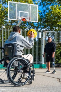 Man in a wheelchair aiming to shoot a basketball while his male friend, who uses a prosthetic leg,