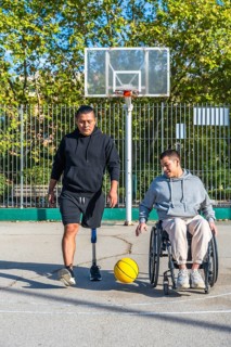 Two friends play basketball on an outdoor court, one man with a prosthetic leg, the other in a