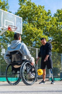 Men with a wheelchair and a prosthetic leg are happily dribbling a yellow basketball on an outdoor