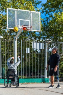 Two men play outdoor basketball, one shooting from a sports wheelchair while the other stands with