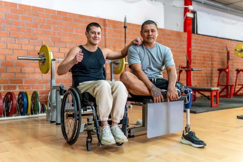 Two adaptive athletes standing together in a gym, one man uses a wheelchair and the other has a