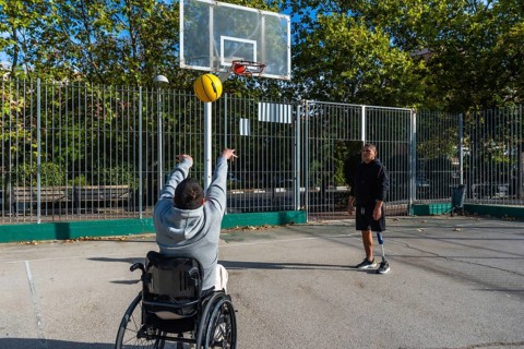 Two young men playing basketball outdoors, one in a wheelchair shooting the ball towards the hoop
