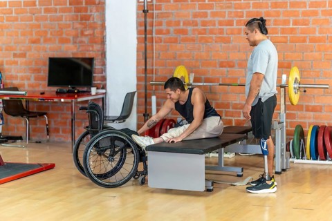 Disabled man in a wheelchair performing stretching exercises on a bench while another man with a