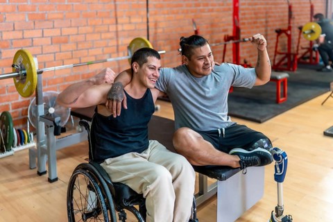 Two smiling men, one using a wheelchair and the other a prosthetic leg, warmly embracing each other