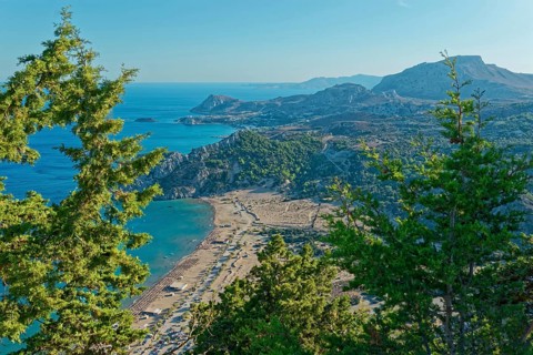 View of the Tsambika sandy beach, with mountains and blue sea, framed by green trees, Church of the