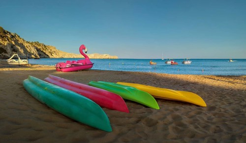Colourful kayaks lying on Tsambika sandy beach, next to a pink flamingo pedal boat, boats floating