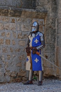 A knight in medieval armour stands with a sword in front of a stone wall, Medieval Town of Rhodes,