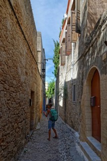 A woman with a rucksack walks through a narrow cobbled alley between historic stone houses,