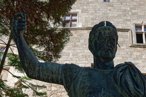 Statue of a man in ancient armour in front of a stone building with windows and trees, Grand