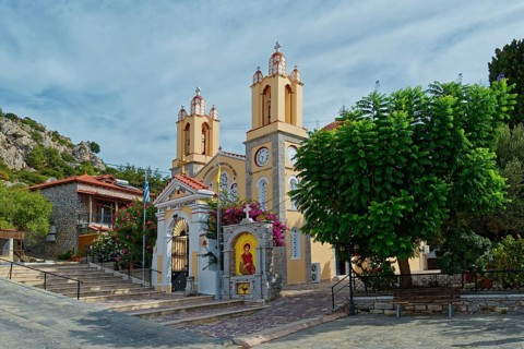 A yellow church with towers and bells surrounded by trees under a cloudy sky, Saint Pantaleon Greek