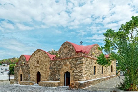 Small stone church Kimisis tis Theotokou with red roofs, surrounded by trees under a clear blue