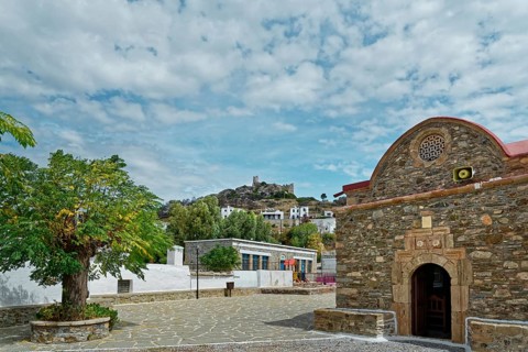 Small church Kimisis tis Theotokou made of stone with red roofs, blue sky and the castle ruins