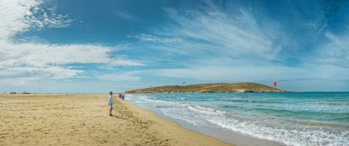 Wide sandy beach of Prasonisi with gentle waves, distant peninsula and kite surfers under a blue