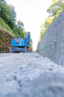 Blue excavator on a path next to a concrete wall with green pipes, Hermann Hessebahn construction