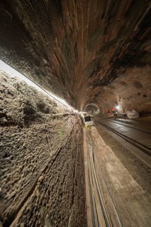 An illuminated tunnel with rails leading into the depths. The walls are made of stone and