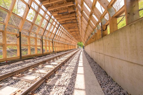 Light-flooded wooden tunnel with railway tracks and concrete path, surrounded by nature,