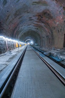 A long, illuminated tunnel with tracks and a deep perspective. The walls appear structured,