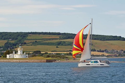A sailing boat with a colourful sail sails near a lighthouse on the coast, Moray Firth, Inverness,