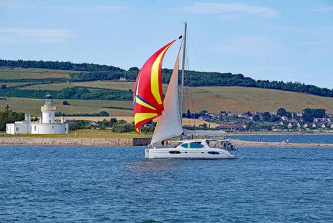 A sailing boat with a coloured sail sails near a lighthouse in front of a green landscape, Moray
