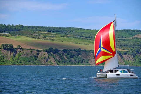 A sailing boat with a vividly coloured flag sails in front of a mountainous coastal landscape,