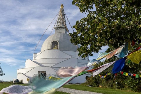 White stupa with colourful prayer flags, peaceful surroundings, green foliage, blue sky, Stupa am