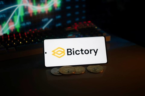 Bictory logo on smartphone amidst coins and stock chart, conveying digital and financial themes