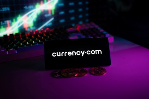 Currency.com logo on phone, with coins and stock chart suggesting financial trading platform