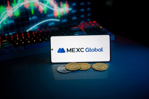 Phone shows MEXC Global logo with crypto coins and trading screen illuminated