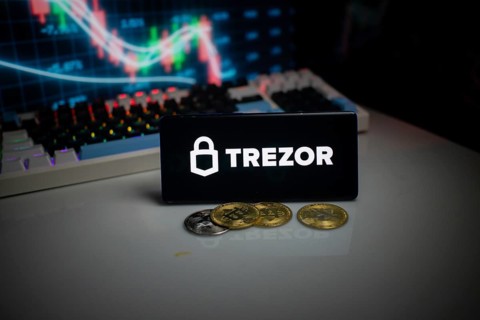 Phone shows Trezor logo with crypto coins and illuminated trading screen