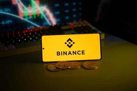 Yellow Binance logo with cryptocurrency coins on a desk, trading graphs in the backdrop